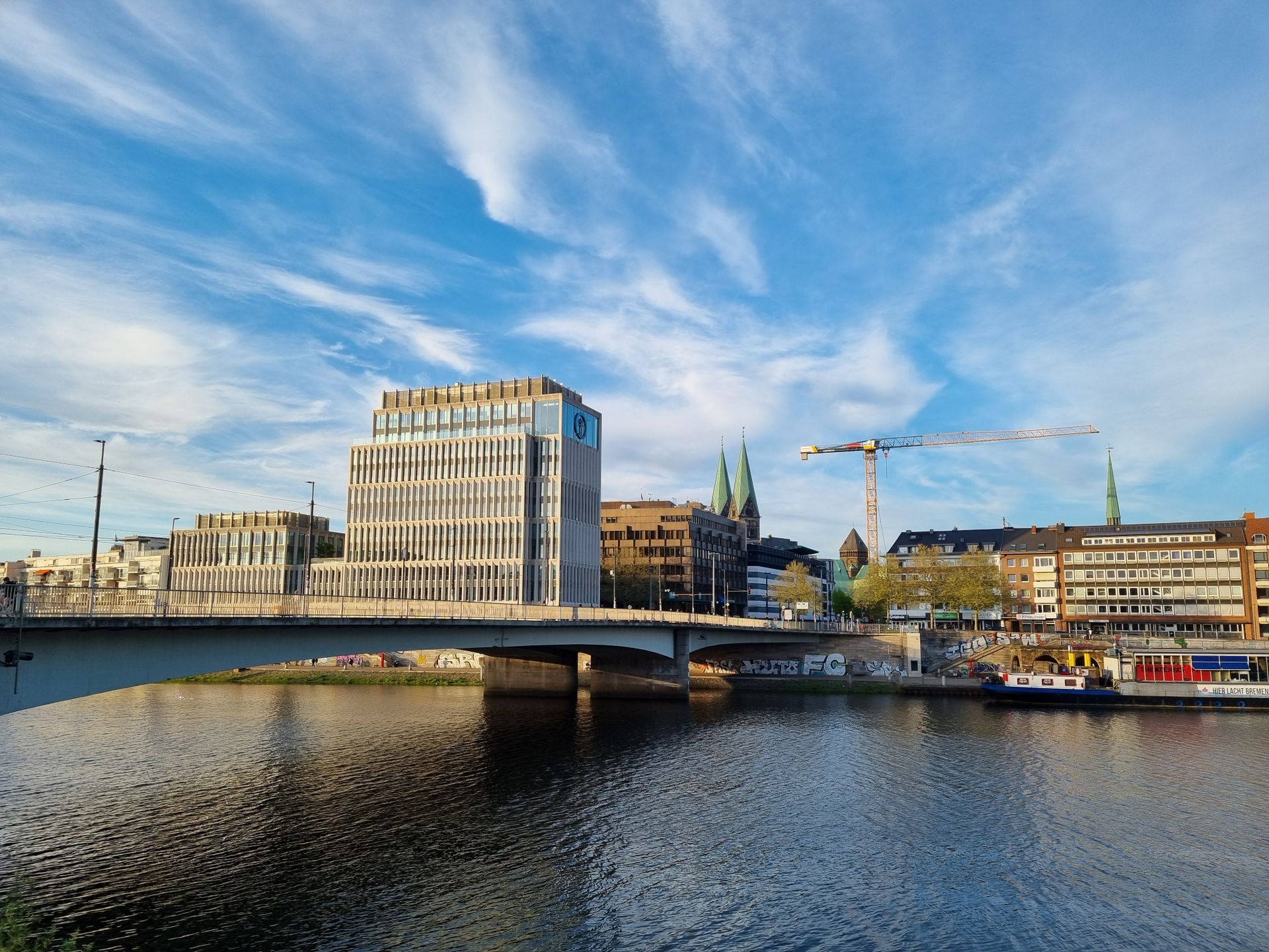 A modern cityscape with a bridge over a calm river, under a bright blue sky.