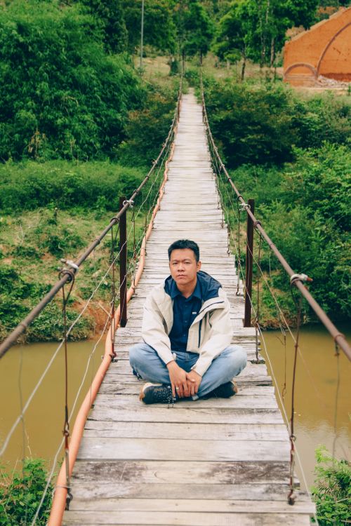 Free Man sitting cross-legged on a suspension bridge in Kon Tum, Vietnam, surrounded by lush greenery. Stock Photo