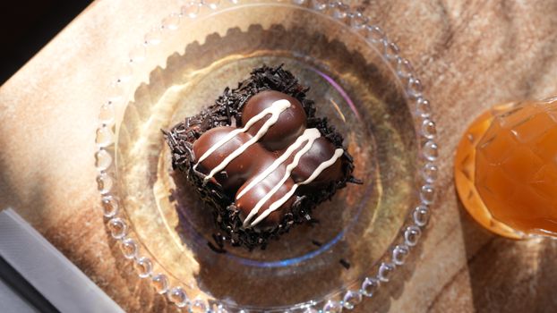 Top view of a chocolate cake with icing on a glass plate, sunlight and a drink nearby.