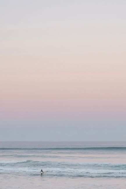 A lone surfer rides the gentle waves under a pastel sunset sky, enjoying the tranquility of the ocean.