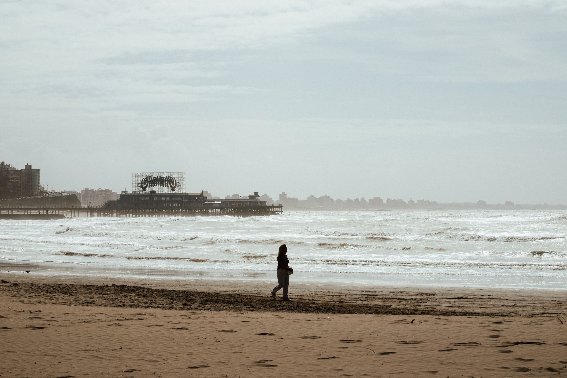 A lone person walks along a deserted beach with a vintage pier in the background.