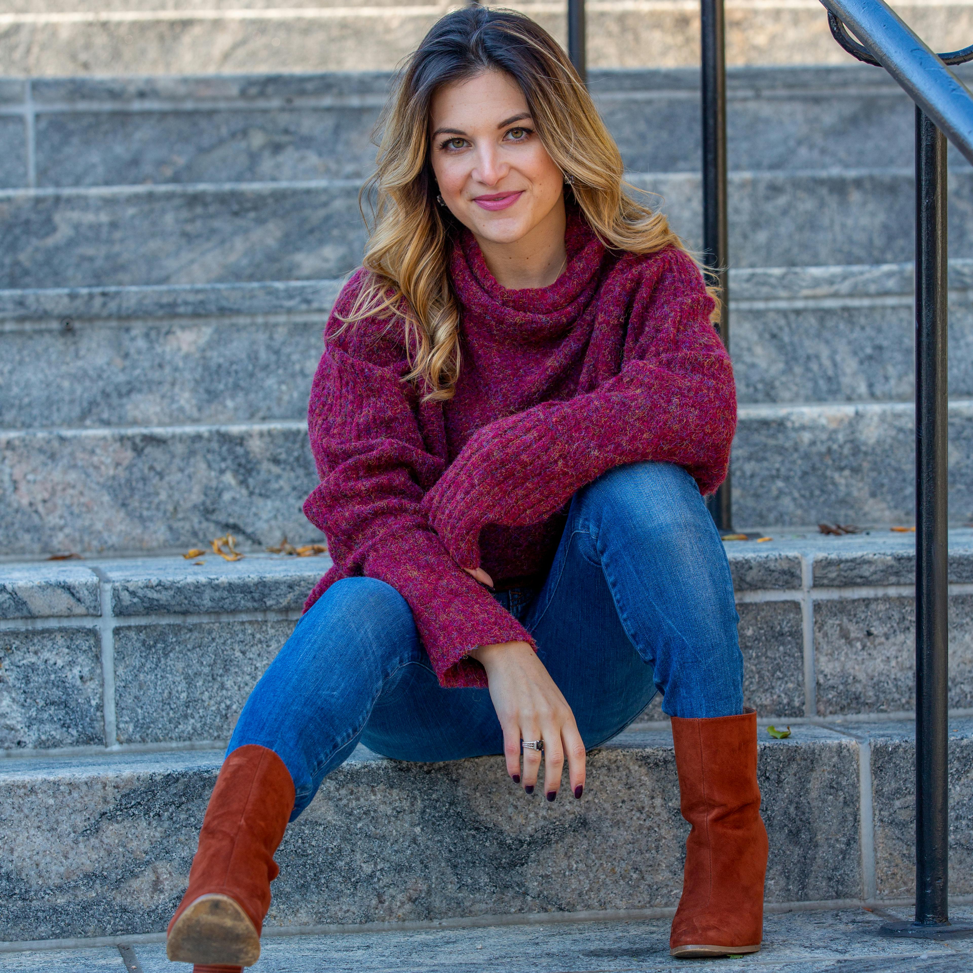 A woman in casual autumn attire smiles while sitting on stone steps in an urban setting.