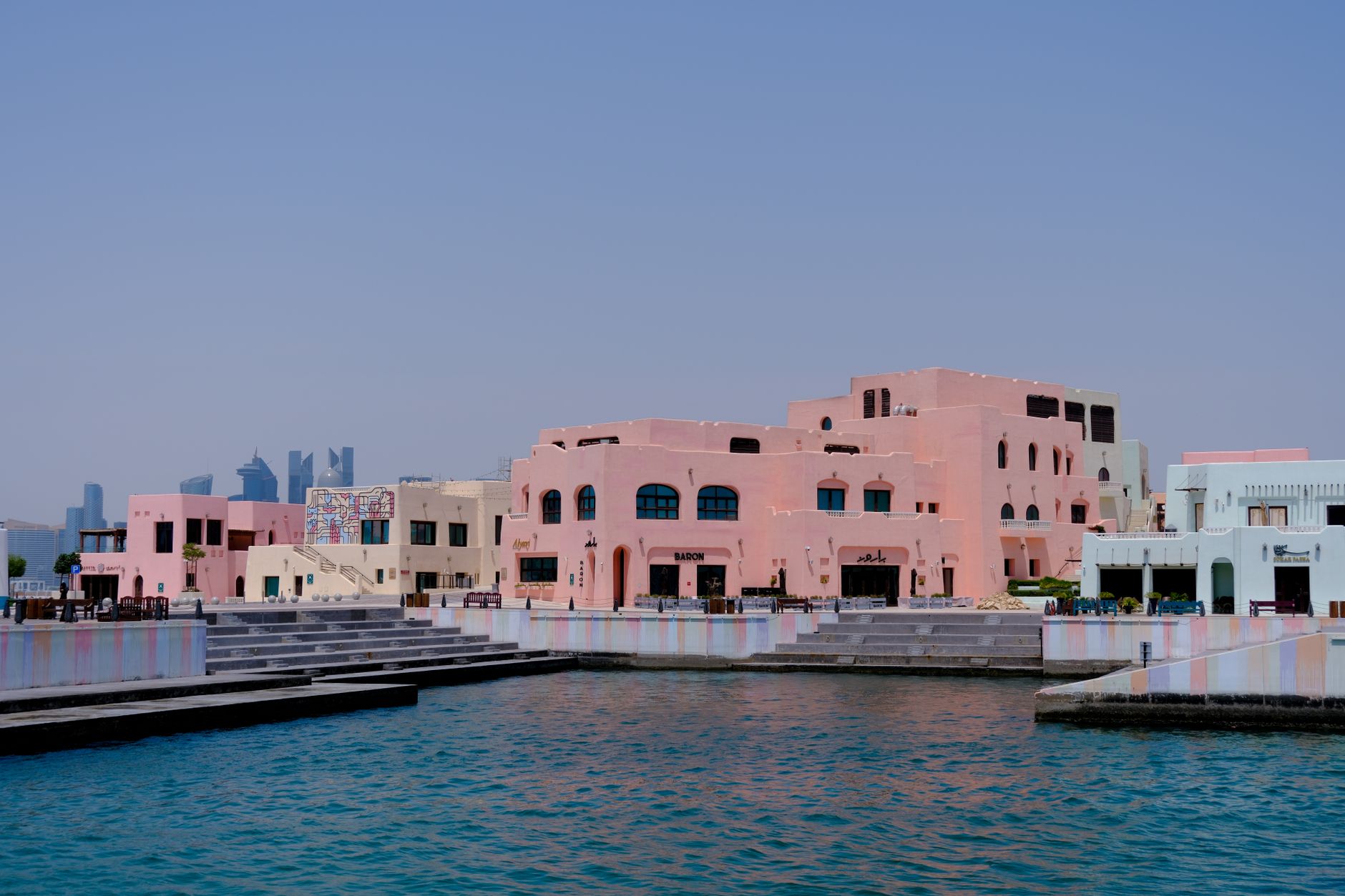 Pastel-colored buildings along a waterfront in Doha, Qatar with a city skyline backdrop.