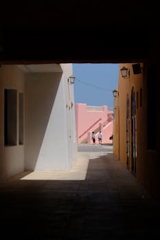 A sunlit alleyway opens to vibrant pastel buildings with people in the distance, captured in daytime.