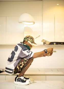 Urban style young man in camo and sneakers posing with floating cash in a modern kitchen.