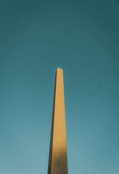 A tall stone obelisk rises prominently against a bright blue sky, showcasing architectural elegance.