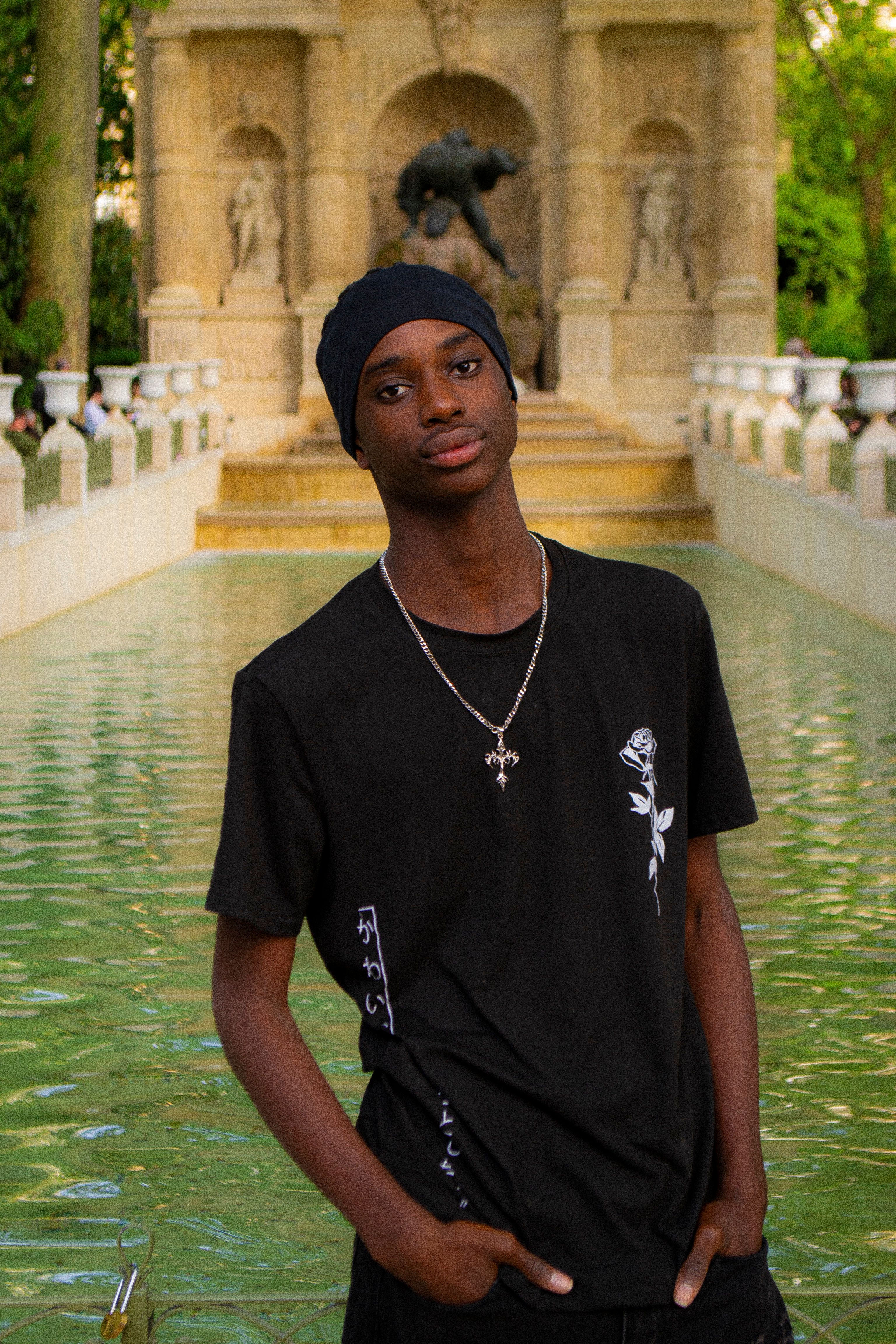 Young man in casual attire posing by a historical fountain in a park, exuding confidence and style.