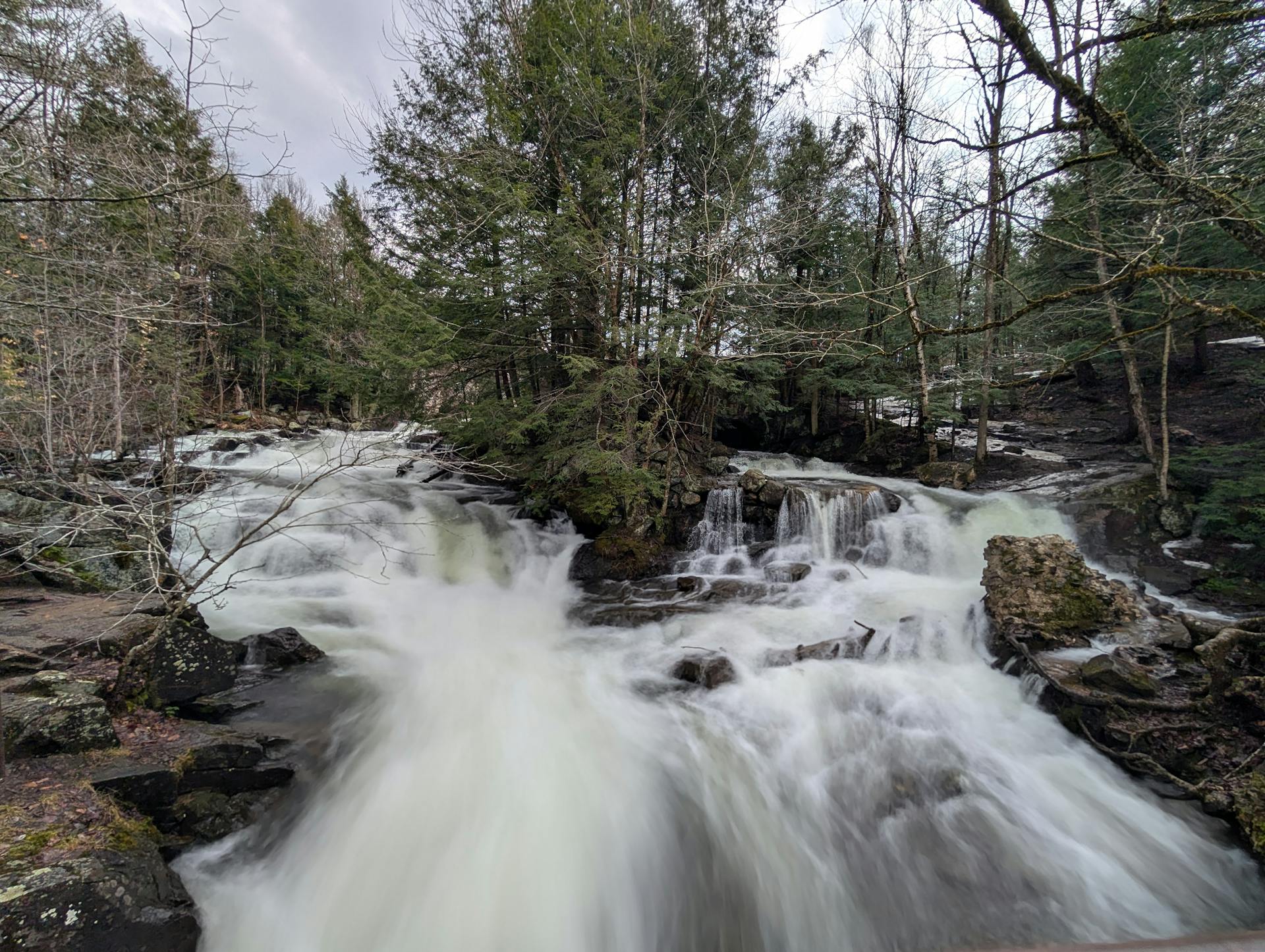 A scenic waterfall cascading through a forested area during early spring, surrounded by evergreen trees.