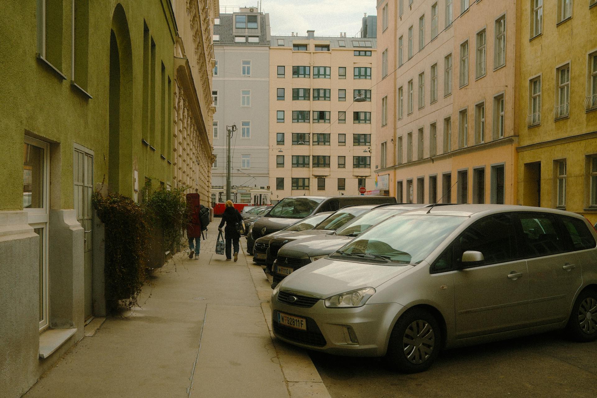 Urban street view with parked cars along colorful buildings, pedestrians walking.