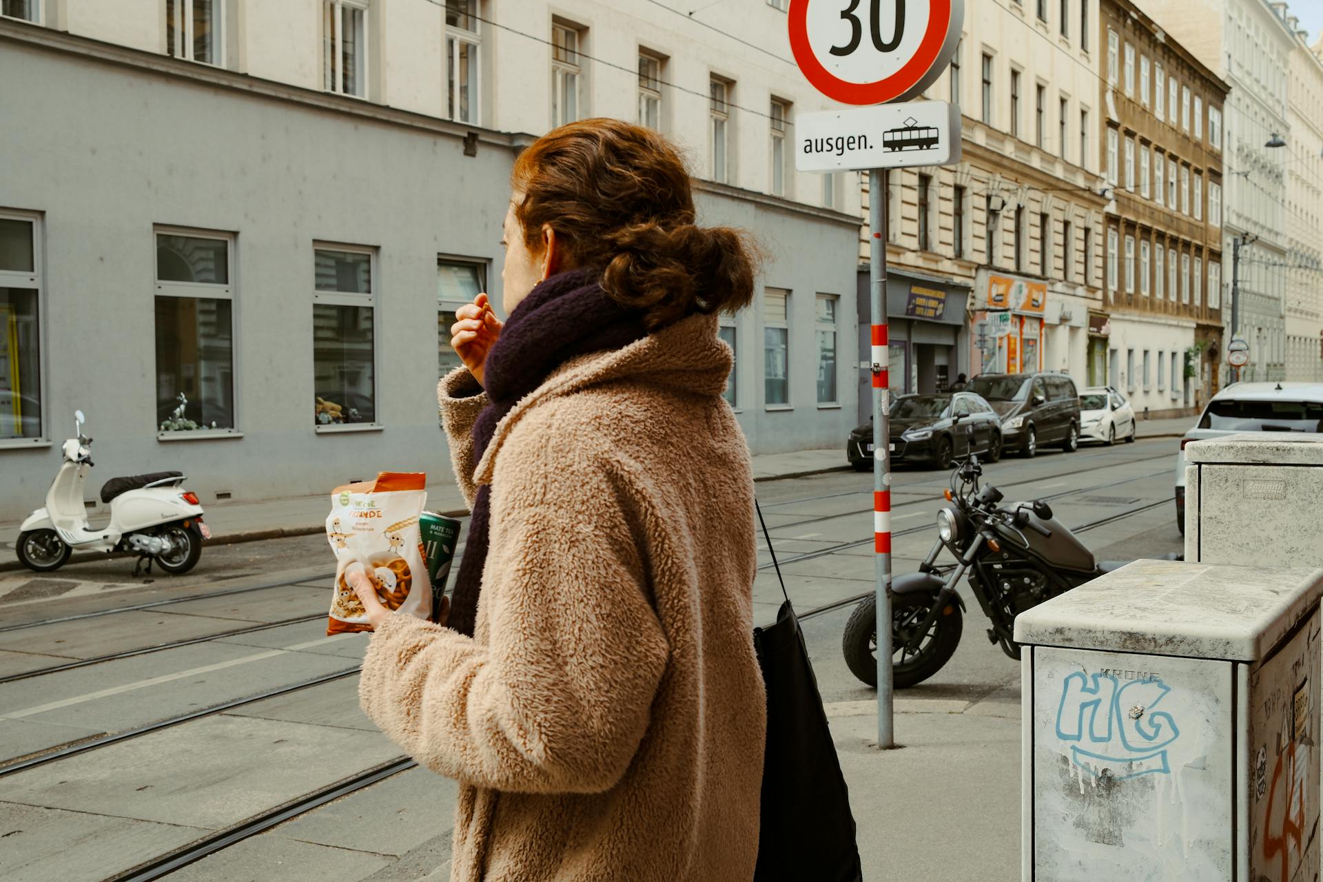 A woman enjoys snacks and coffee while walking through a European city street, showcasing urban life.