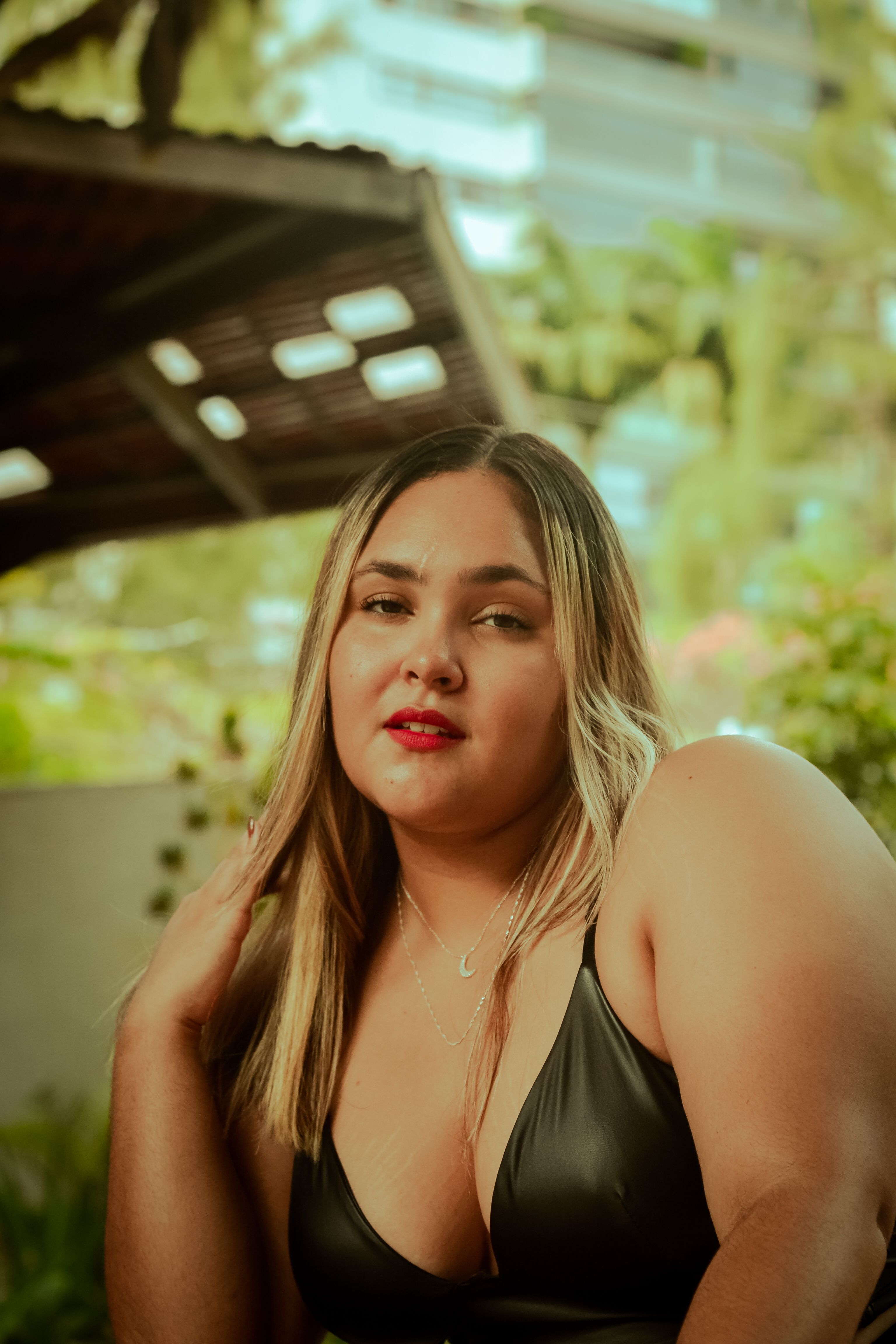 A confident woman poses outdoors in a black top, surrounded by greenery and modern architecture.