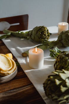 Warm sunlight illuminates a rustic table with artichokes and a lit candle, creating a cozy atmosphere.