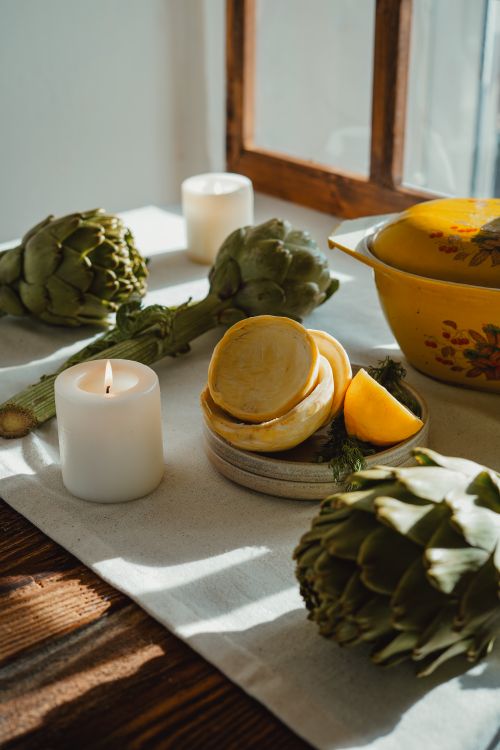 Free Sunlit rustic table setting with artichokes, lemon, and candle for cozy ambiance. Stock Photo