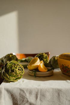 A rustic kitchen scene with artichokes, cut lemons, and a decorative bowl under natural sunlight.