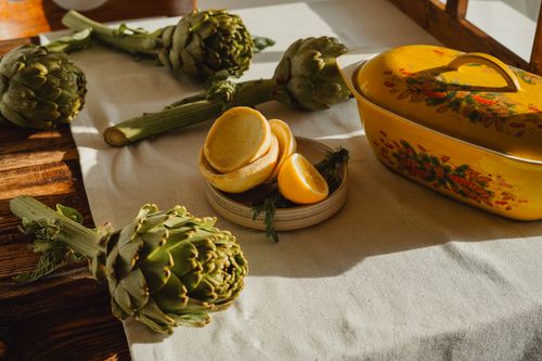 Free Artichokes and sliced lemons with a vintage yellow casserole dish. Sunlit rustic kitchen scene. Stock Photo