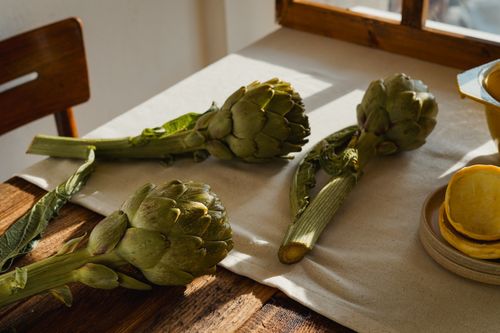 Free Three fresh artichokes on a rustic wooden table with sunlit ambiance. Perfect for a culinary setup or kitchen decor. Stock Photo