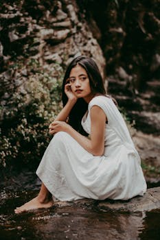 A woman in a white dress sitting by the water, exuding natural beauty and calmness.