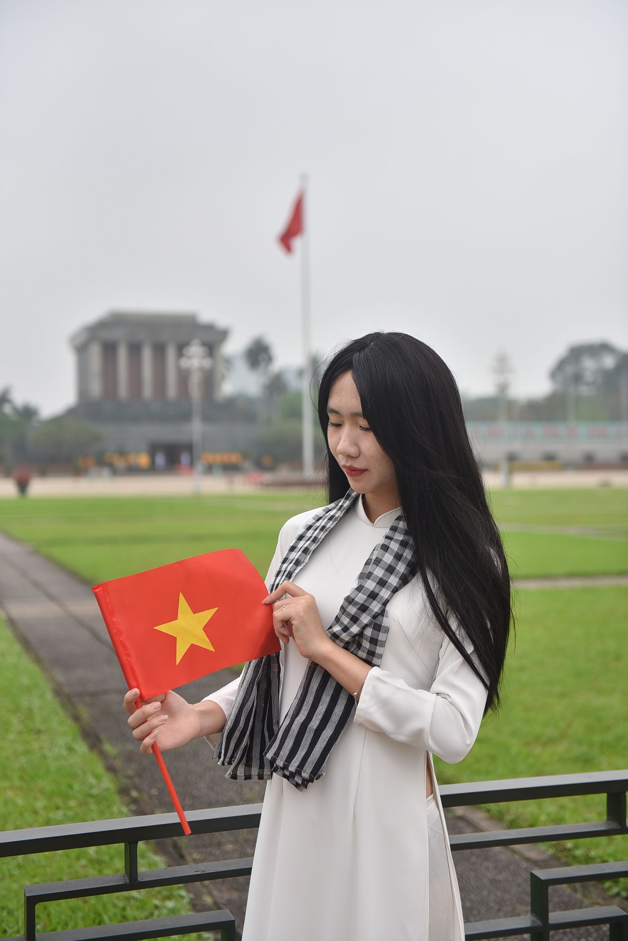 A young woman in traditional attire holding a Vietnamese flag in front of a monument in Hanoi.