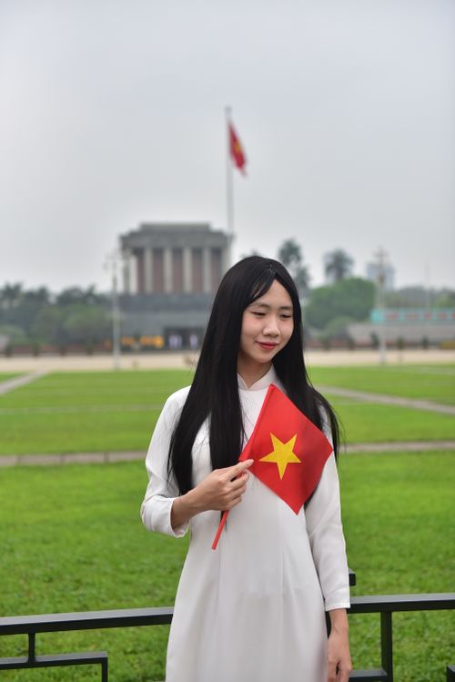 Free Young woman in traditional attire holding Vietnamese flag near famous mausoleum in Hanoi. Stock Photo