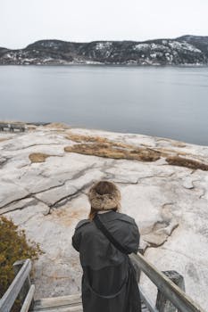 Person stands on a rocky ledge overlooking a serene body of water and distant snowy hills.