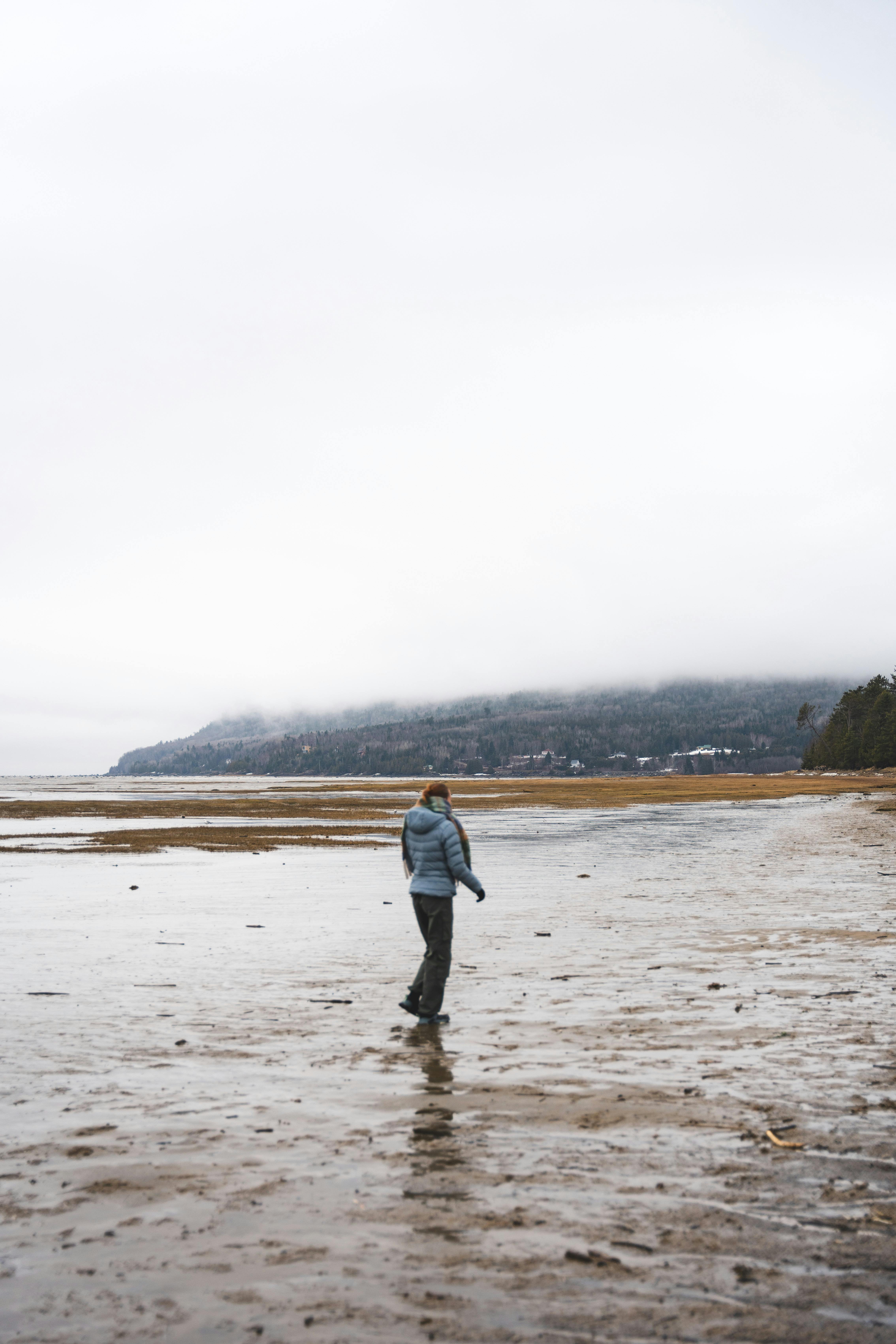A solitary figure walks along a misty beach in Charlevoix, Quebec, with distant mountains shrouded in clouds.