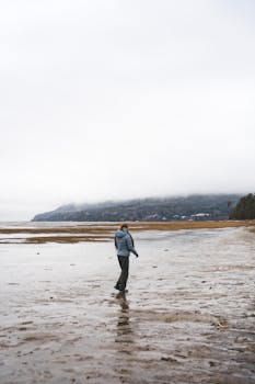 A solitary figure walks along a misty beach in Charlevoix, Quebec, with distant mountains shrouded in clouds.