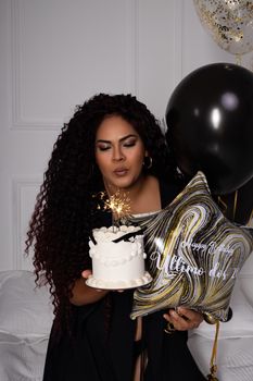 Woman holding birthday cake with sparklers and balloons indoors.