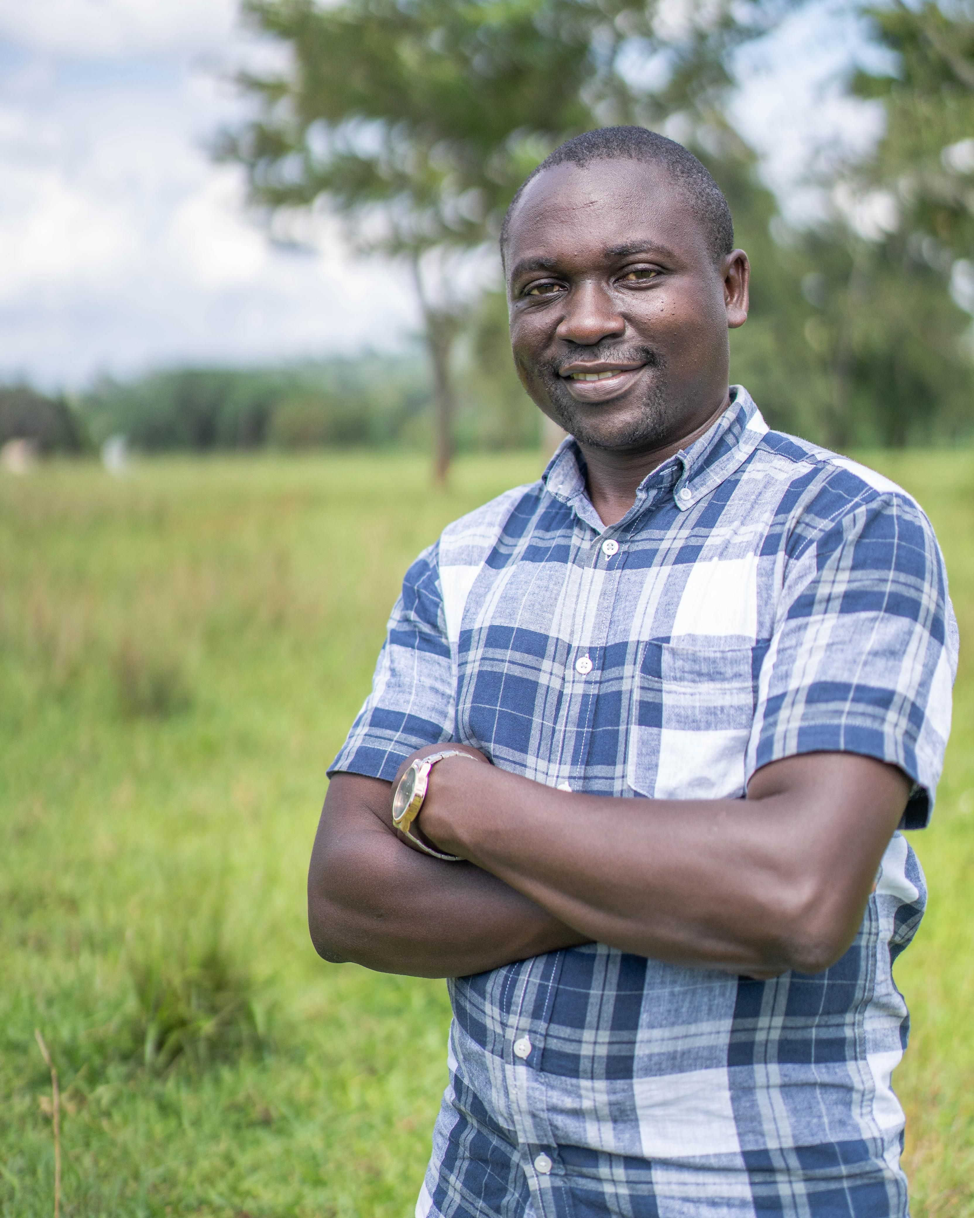 Smiling man standing confidently in a green field with trees in the background.