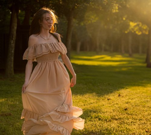 Free Young woman in a flowing peach dress walks gracefully in a sunlit park during sunset. Stock Photo