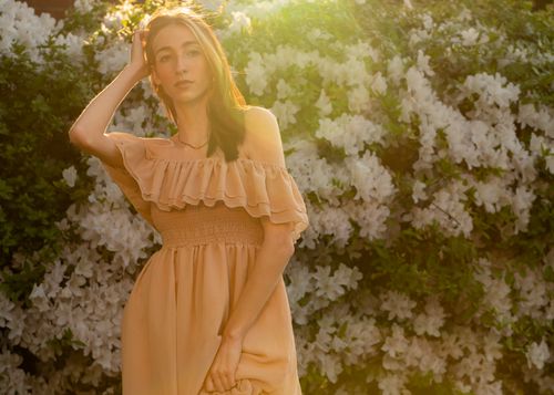 Free A young woman in a peach dress poses outdoors with blooming white flowers, illuminated by sunlight. Stock Photo