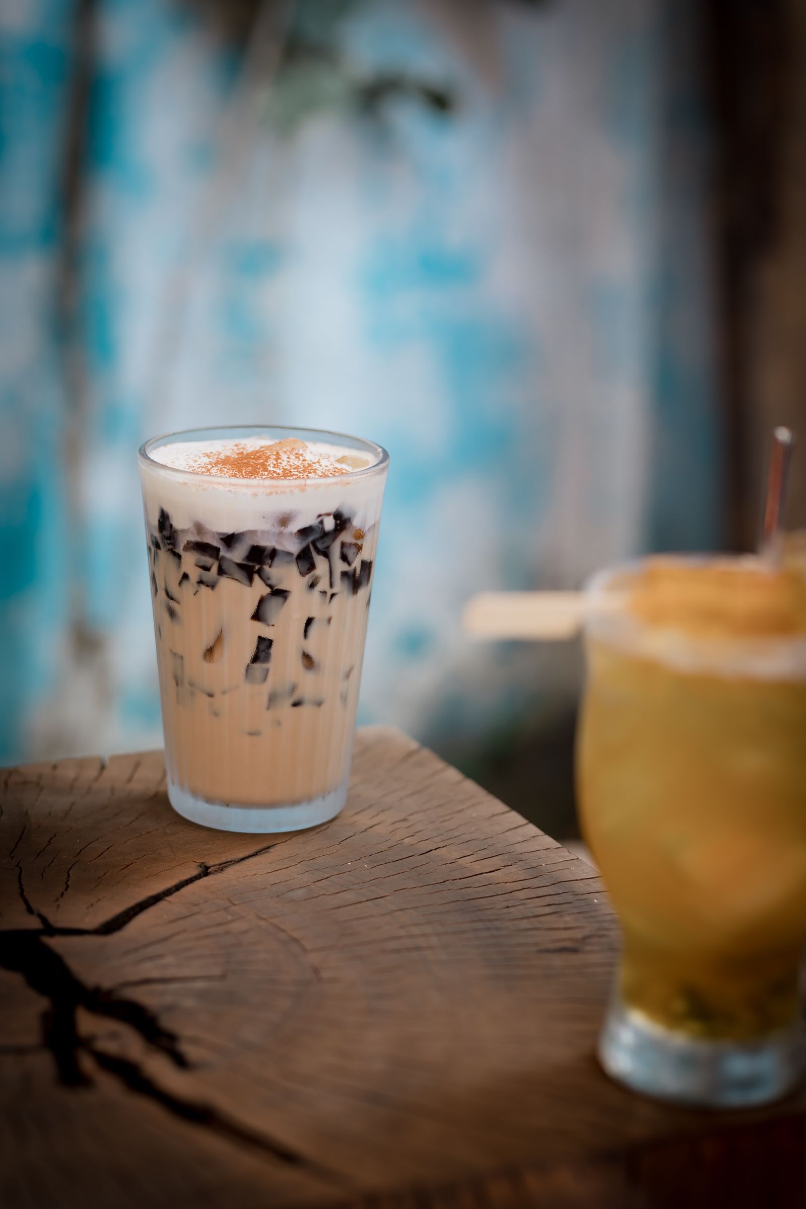 A glass of creamy milk tea with grass jelly in focus next to a glass of clear golden fruit tea on a wooden table