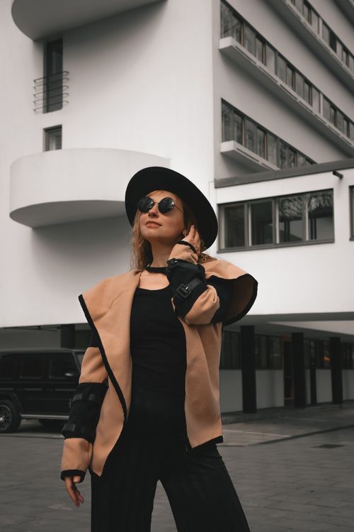 Free Stylish woman wearing hat and sunglasses poses confidently in front of a modern building. Stock Photo