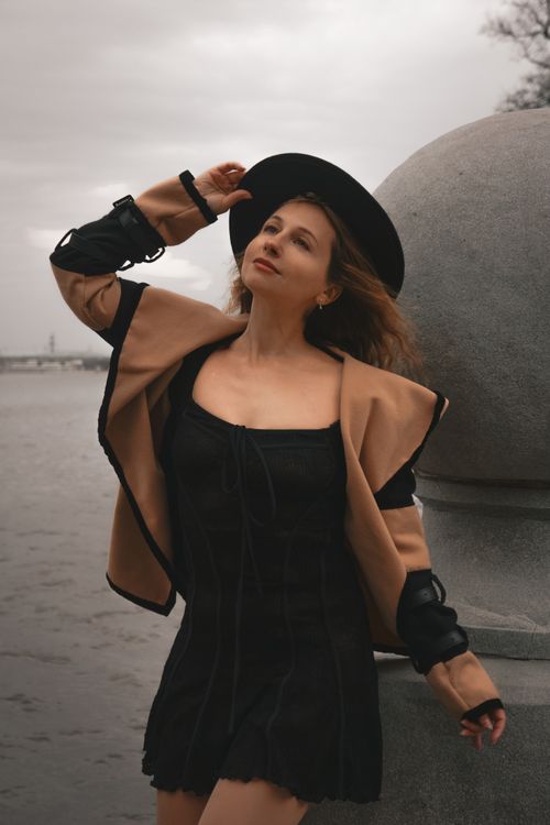 Free Stylish woman in black dress and hat striking a pose by a riverside in an autumn setting. Stock Photo