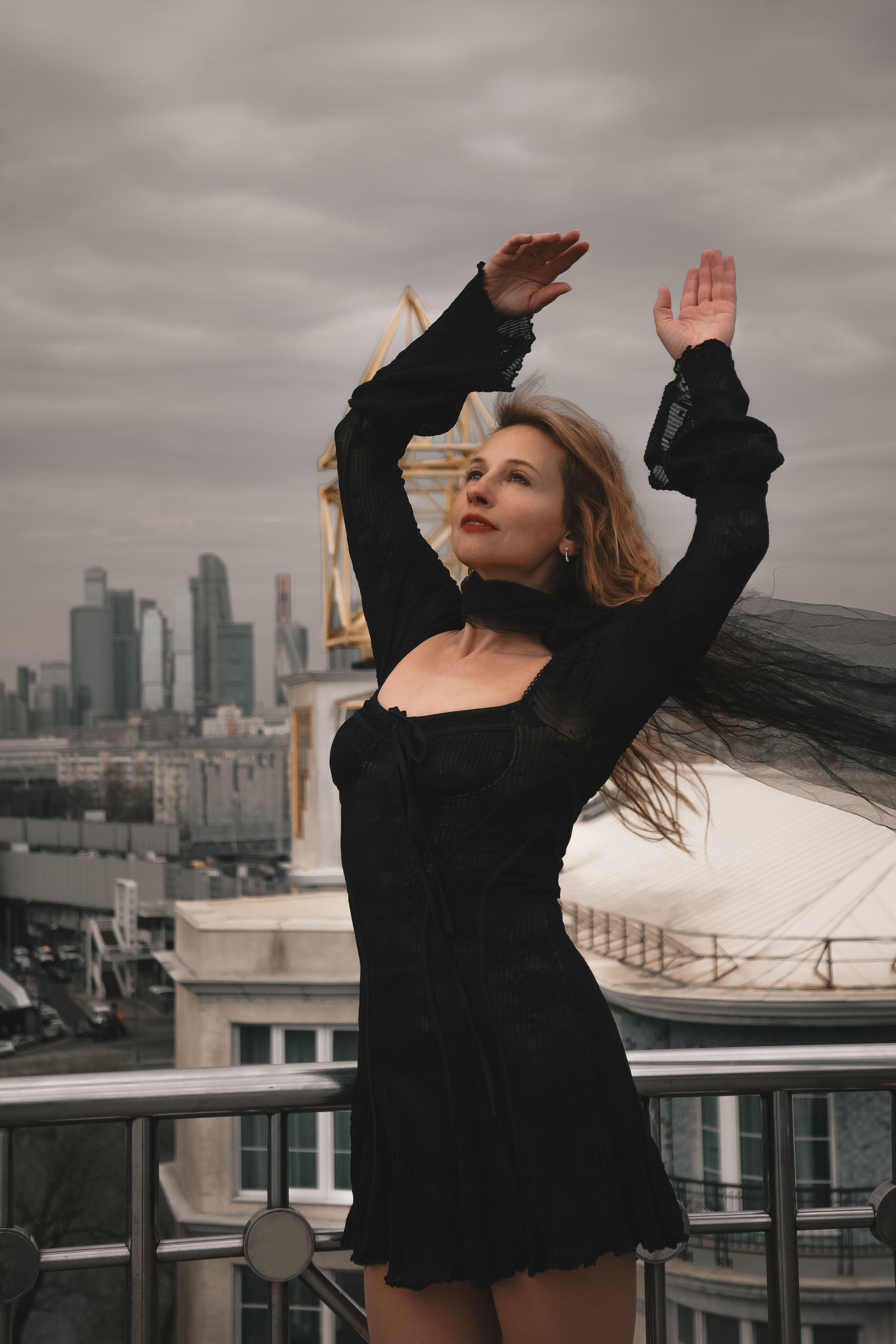 Elegant woman in a black dress striking a pose on a rooftop with cityscape in the background.