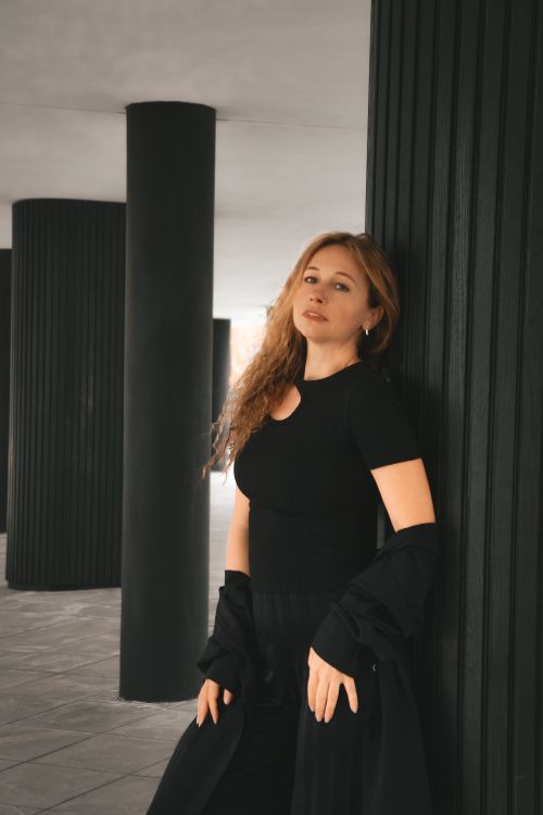 Free A fashionable woman leans against dark columns in a modern architectural setting. Stock Photo