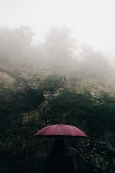 A person stands under a red umbrella facing a misty mountain landscape.