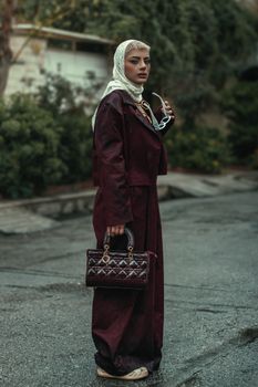 Elegant woman with headscarf holding sunglasses and handbag outdoors.