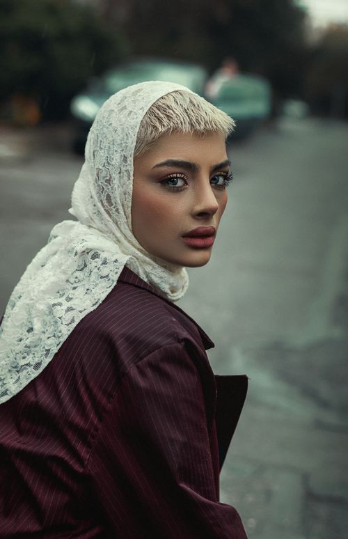 Free A fashionable woman wearing a lace headscarf and coat on a city street, captured in a moody style. Stock Photo