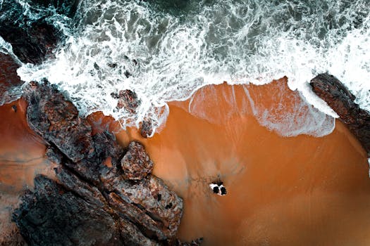 A stunning aerial shot capturing the interplay between rocky shores and ocean waves crashing on the beach.