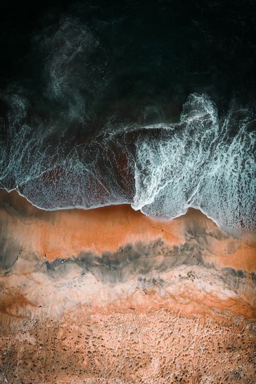 Free A stunning aerial shot capturing the dramatic contrast of ocean waves crashing onto a sandy beach shoreline. Stock Photo
