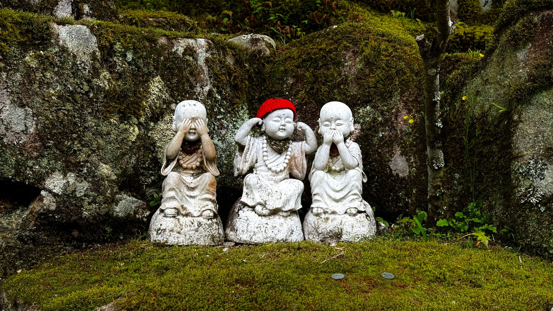 Three stone monk statues symbolize 'see no evil, hear no evil, speak no evil' in a lush, mossy garden.