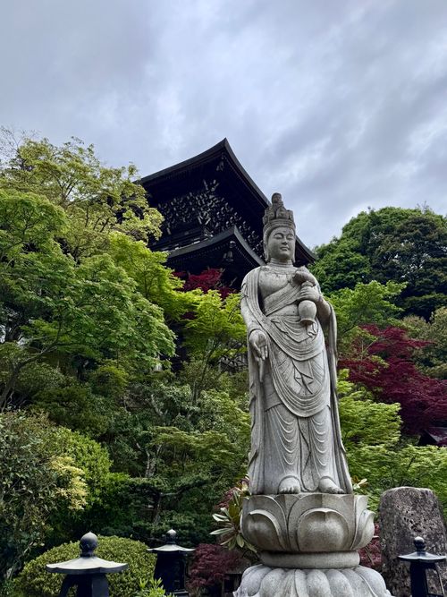 Free Stone statue of Buddhist figure in lush green Japanese garden with temple in background. Stock Photo