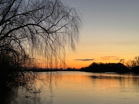 A tranquil sunset view over a calm river with silhouetted trees and a vivid orange sky.