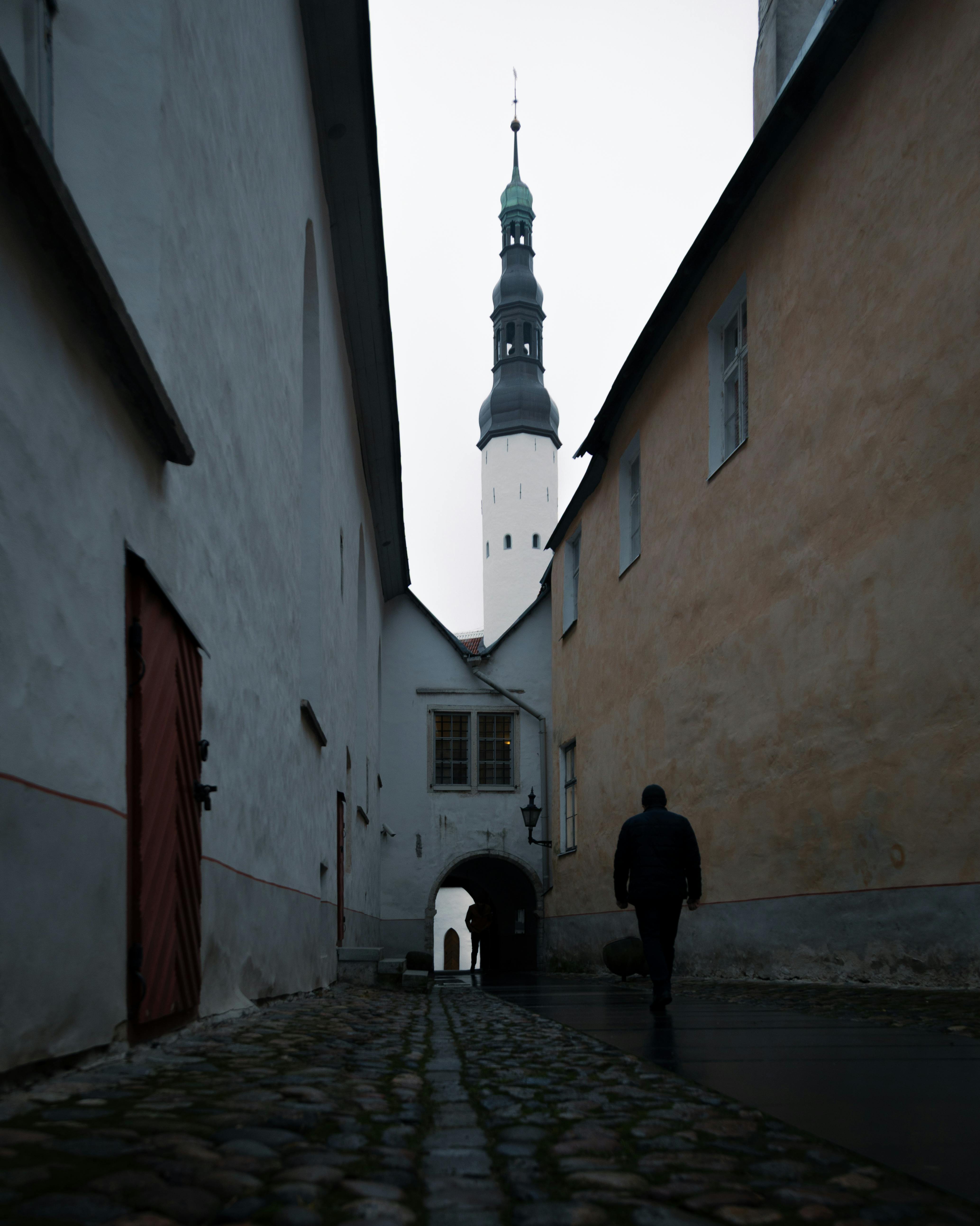 A solitary man walks through a narrow street in Tallinn, Estonia's Old Town, marked by medieval architecture and a prominent spire.