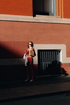 A stylish woman enjoys a sunny day in Soho, New York City, holding coffee and standing by a red brick wall.