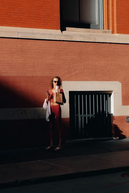 A stylish woman enjoys a sunny day in Soho, New York City, holding coffee and standing by a red brick wall.