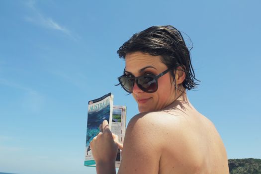 Woman with sunglasses reading a travel guide on a sunny Australian beach.