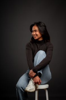Smiling woman in jeans and black top, seated casually on stool in studio setting.