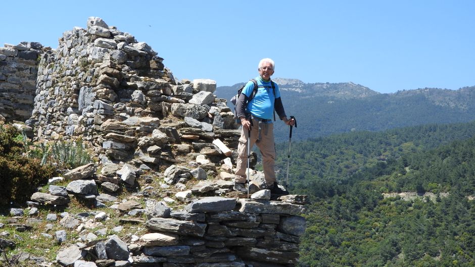A senior man hikes among ancient ruins with trekking poles under a clear blue sky.