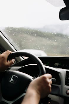 Close-up of hands on steering wheel in a car during rainy day drive.